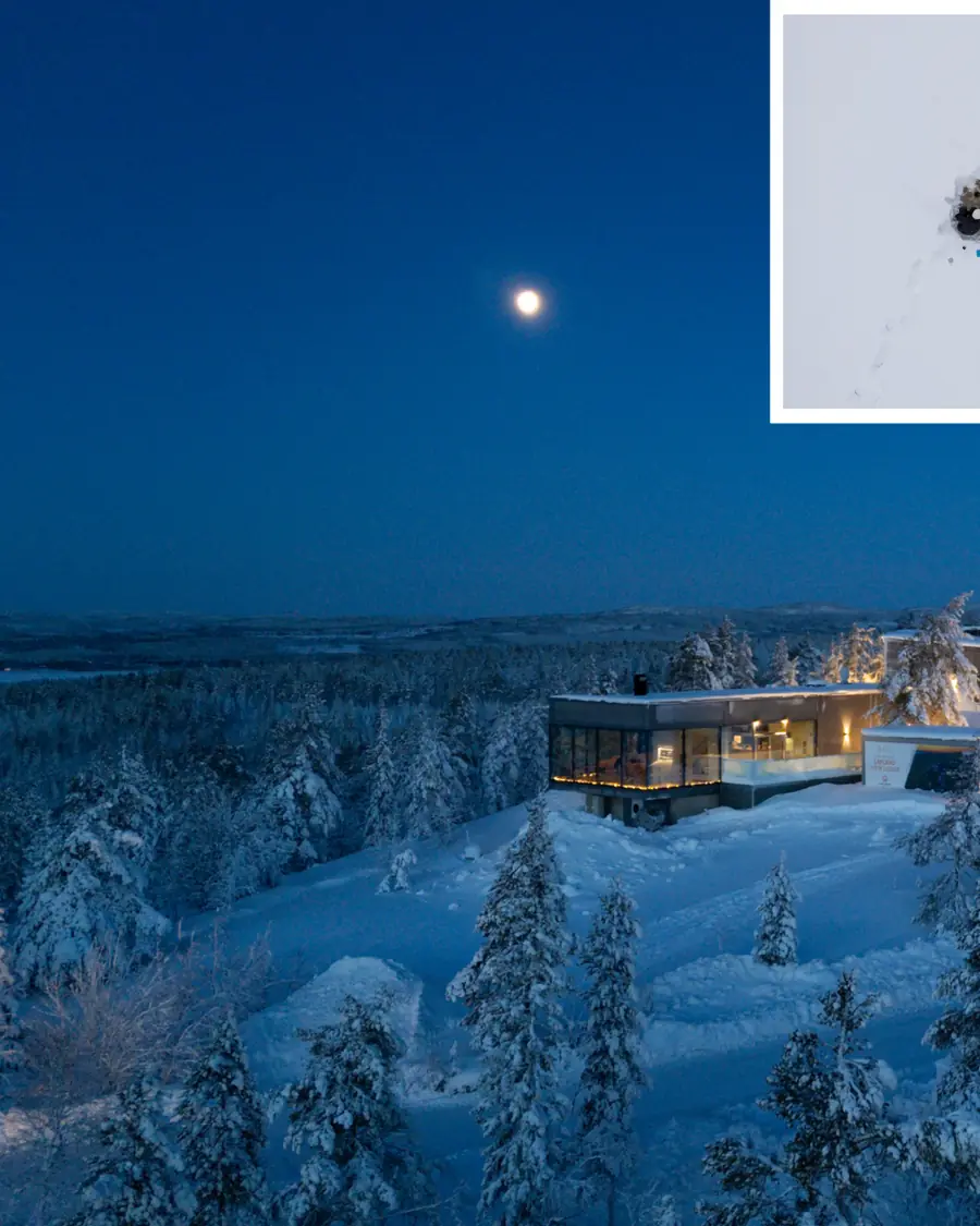 A glass lodge lit from within on a snowy ridge under a full moon at blue hour