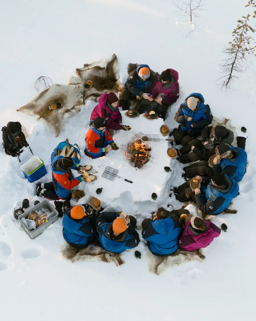 A group eating lunch on reindeer hides around a fire, seen from above